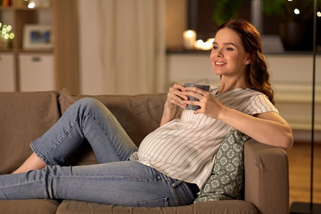 母亲喝茶摄影照片_怀孕和人的概念—愉快的微笑孕妇喝茶在家里的沙发上。微笑的孕妇喝茶在家
