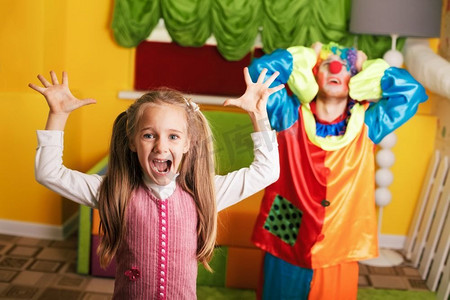 小丑背景摄影照片_小女孩在生日聚会上玩心烦意乱的小丑。彩色沙发在背景上。小女孩玩心烦意乱的小丑。