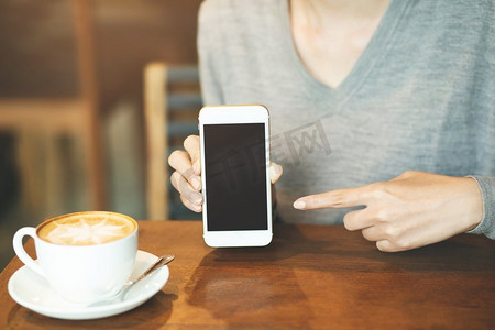智能电话摄影照片_妇女手拿着移动智能电话与空白桌面屏幕和手指接触，而喝咖啡在咖啡店。	