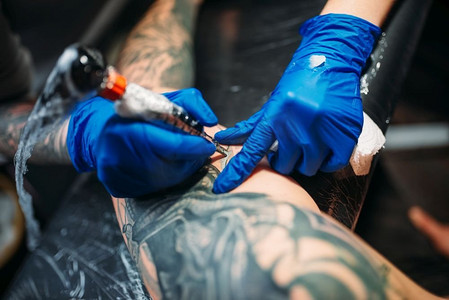 纹身师摄影照片_女纹身师用机器在男性肩膀上纹身。专业纹身在沙龙。纹身师通过机器在男性肩膀上纹身