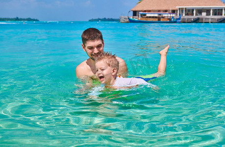 全家一起去旅行摄影照片_三岁的学步男孩和父亲一起学习游泳。暑假全家去马尔代夫度假。