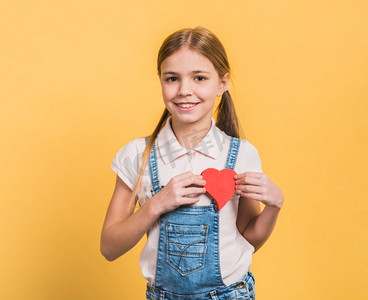 肖像微笑的女孩显示红色纸切出心形反对黄色背景