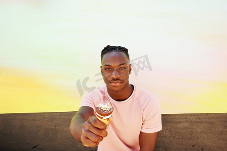 年轻英俊的黑人男子穿着一件粉色t恤，手持冰激凌蛋卷，在阳光明媚的夏天，在一面画好的墙上吃着。