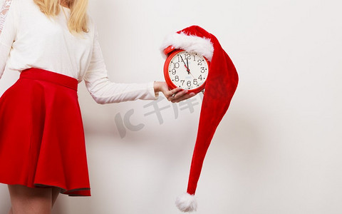 戴着圣诞老人帽子的女人拿着闹钟的特写。圣诞时节概念..拿着闹钟的女人。圣诞节到了。