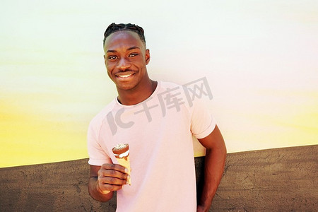 一个年轻英俊的黑人男子穿着粉红色的T恤，微笑着，在夏天的日出或晴天的墙上拿着一个冰淇淋甜筒