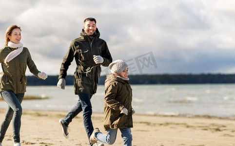 海滩跑步摄影照片_家庭、休闲、人的概念-快乐的父母和小女儿沿着秋天的海滩奔跑。幸福的一家人沿着秋天的海滩跑步