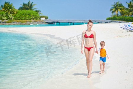 全家一起去旅行摄影照片_一个三岁的蹒跚学步的男孩和妈妈一起走在海滩上。暑假全家去马尔代夫度假。
