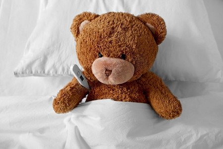 积木玩具卡头摄影照片_医学，保健和儿童概念—生病的泰迪熊玩具头与温度计躺在床上。生病的泰迪熊玩具头与温度计在床上