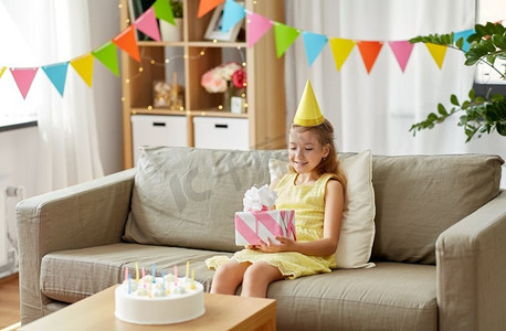 童年和庆祝概念--戴着派对帽的快乐女孩，家里有生日礼物和蛋糕。戴着派对帽的快乐女孩在家里带着生日礼物