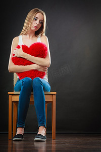 心碎摄影照片_心碎的爱情概念。悲伤的不愉快的女人坐在椅子拥抱红心枕头黑暗背景