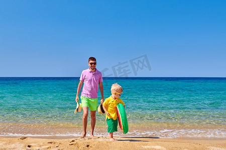 爸爸拿摄影照片_两岁的蹒跚学步的男孩在海滩上与父亲走，拿着充气戒指。暑假家庭度假。希腊，西托尼亚。 