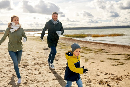 家庭、休闲、人的概念-快乐的父母和小儿子沿着秋天的海滩奔跑。幸福的一家人沿着秋天的海滩跑步