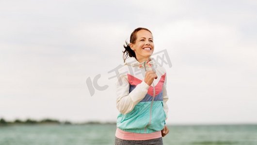 健身，运动和健康的生活方式概念—微笑的女人沿着海滩跑步。微笑的女人沿着海滩奔跑
