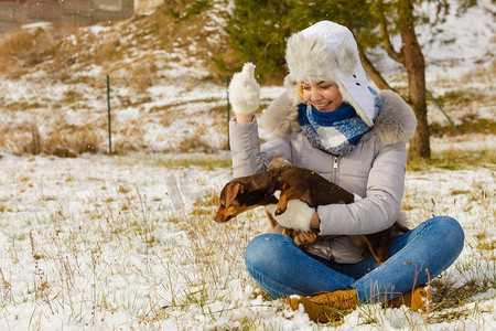 下雪玩耍摄影照片_年轻女子在外面的雪地里玩得很开心。一只母狗在寒冷的冬日和她的纯种小狗玩耍。一名女子在冬天与狗玩耍