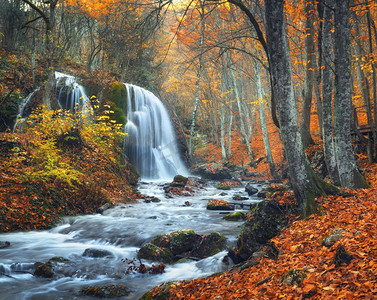 红色和橙色的树叶摄影照片_美丽的瀑布在山河在五颜六色的秋天森林与红色和橙色的叶子在日落。自然景观