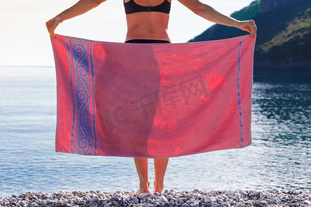妇女覆盖她的臀部与粉红色毛巾站在海岸线放松。假期和旅行概念妇女在海滩覆盖臀部用毛巾