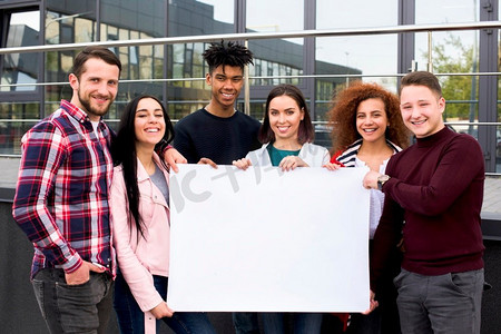 微笑的多民族学生举着空白的白色海报站在玻璃建筑前