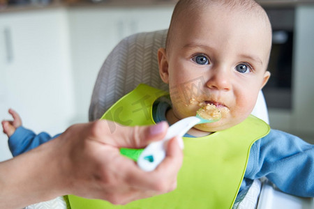 微笑的婴儿男孩的肖像在家里高椅子被喂固体食物由母亲用勺子                             