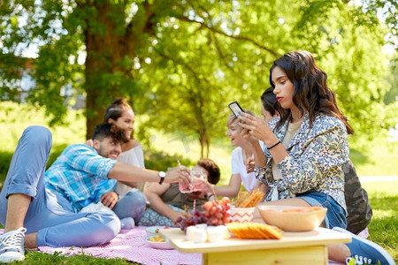 休闲，技术和人的概念—妇女使用智能手机在野餐与朋友在夏天公园。妇女使用智能手机在野餐与朋友