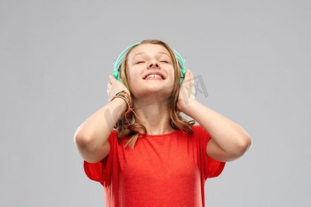 音乐，科技和人的概念-戴着耳机，穿着灰色背景下的红色T恤的快乐少女。戴着耳机的快乐少女
