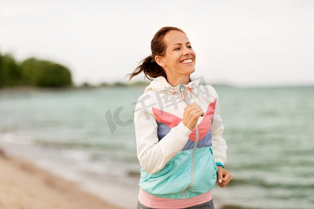 健身，运动和健康的生活方式概念—微笑的女人沿着海滩跑步。微笑的女人沿着海滩奔跑