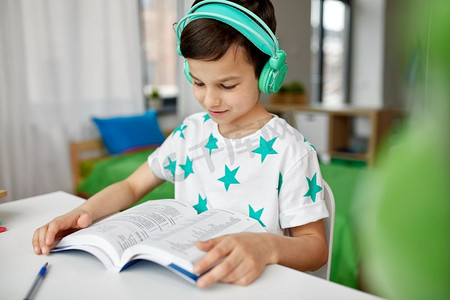 教育、童年与科技理念--快乐男生戴着耳机在家看书。戴着耳机在家学习课本的男孩
