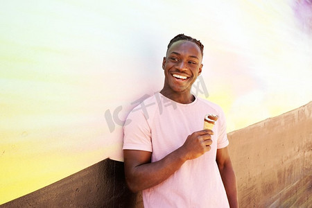 一个年轻英俊的黑人男子穿着粉红色的T恤，微笑着，在夏天的日出或晴天的墙上拿着一个冰淇淋甜筒
