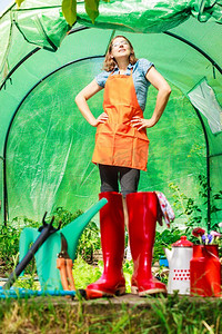 园艺工具放在室外花园里，红色胶靴可以冲水。背景中在温室里工作的女农民