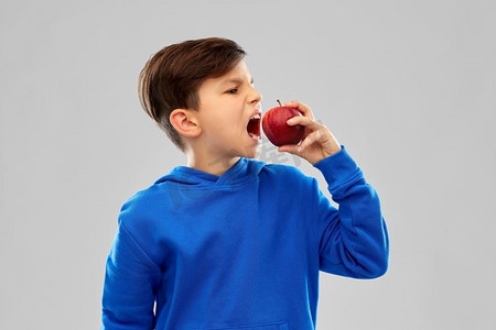 抱着东西的男孩摄影照片_食物，饮食和健康饮食理念-穿着蓝色连帽衫的愤怒男孩，灰色背景上有红色的苹果。穿着蓝色连帽衫、戴着红苹果的愤怒男孩