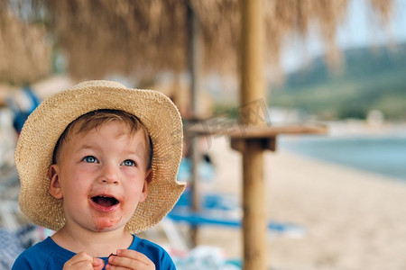 海滩伞摄影照片_2岁的小男孩在海滩上吃水果。快乐的孩子肮脏的脸。