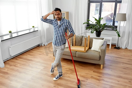 清洁、家务和家政概念—疲惫的印度男子与扫帚扫地在家里。男人用扫帚打扫地板