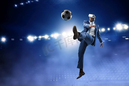 黑人男子与虚拟现实头盔踢足球在夜间体育场与燃烧的灯光。黑人商人在虚拟现实足球比赛