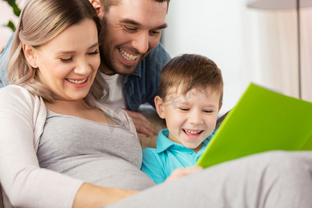 怀孕，人和家庭的概念—快乐怀孕的母亲，父亲和小儿子在家里的沙发上看书。快乐的家庭阅读书在家里