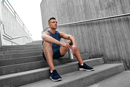 健身，运动和人的概念—疲惫的年轻人与瓶装水坐在楼梯上。疲惫的运动员拿着瓶子坐在楼梯上