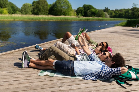 户外喝啤酒摄影照片_休闲、野餐和人的概念-朋友们在夏季公园的湖畔码头喝啤酒和苹果酒。朋友们在湖边码头喝着啤酒和苹果酒