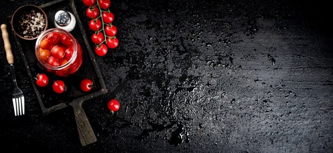 把腌制的西红柿放在切菜板上的玻璃瓶里。在黑色背景上。高质量的照片。把腌制的西红柿放在切菜板上的玻璃瓶里。