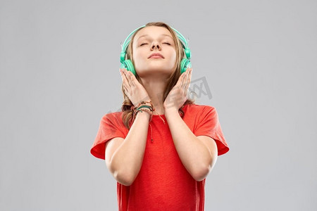 音乐，科技和人的概念-戴着耳机，穿着灰色背景下的红色T恤的快乐少女。戴着耳机的快乐少女