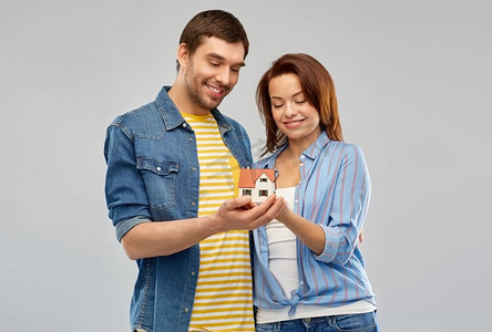 房地产、家庭和抵押概念—微笑的夫妇持有房子模型在灰色背景。微笑的夫妇举行房子模型