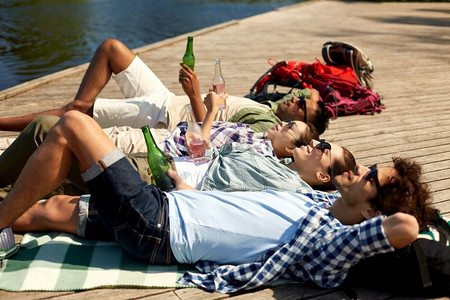 休闲、野餐和人的概念-朋友们在夏季公园的湖畔码头喝啤酒和苹果酒。朋友们在湖边码头喝着啤酒和苹果酒