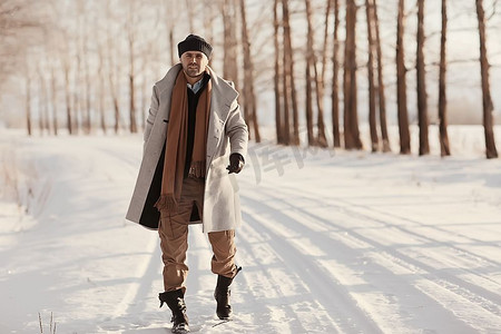 冬季风格摄影照片_穿着大衣/冬季风格的时髦男子，在冬季风景的背景下行走，下雪的天气，保暖的衣服