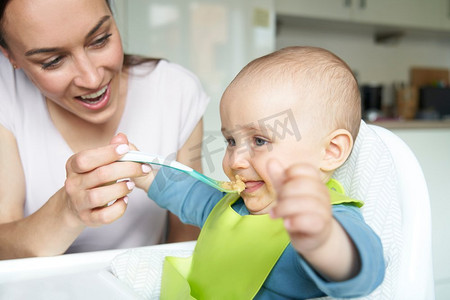 微笑的8个月大的男婴在家里在高脚椅被喂固体食物由母亲用勺子    