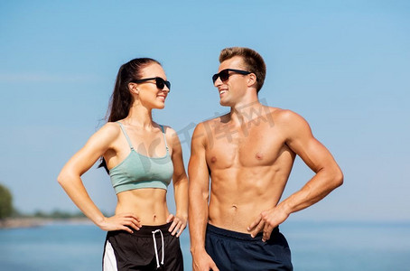 健身，运动和生活方式的概念—快乐的夫妇在运动服和太阳镜在海滩。快乐的夫妇在运动衣服和阴影在海滩上