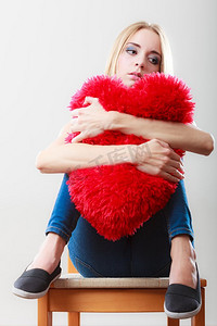 心碎的爱情观。伤心不开心的女人坐在椅子上抱着红心枕头