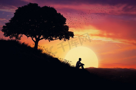 一个男孩独自坐在大自然中心的一座小山上，看着落日的背景。远离人群，等待大自然的治愈力量。