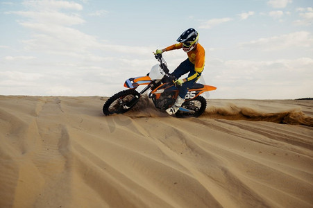 骑摩托的男人摄影照片_熟练的专业摩托车手进行特技表演。摩托车手在沙滩上滑行时保持平衡。骑摩托车的人在滑沙上做特技