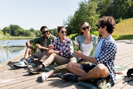 休闲，野餐和人的概念—朋友喝啤酒和苹果酒在湖码头在夏季公园。朋友们在湖码头喝啤酒和苹果酒