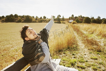 一个年轻人与伸展的手臂和温暖的围巾享受早晨秋天的阳光在一个黄色领域与背光从蓝天