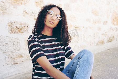 一个严重的年轻黑人妇女的肖像戴眼镜，牛仔裤和条纹t恤，坐在地上和卷发