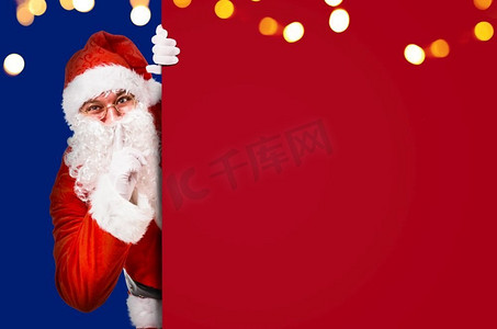 服装要求摄影照片_圣诞老人用手指放在嘴唇上，要求安静，上面有五颜六色的广告牌和文案空间。圣诞老人要求保持沉默