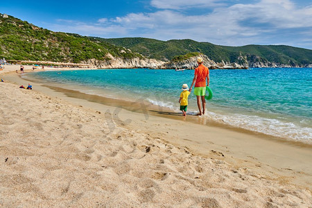 爸爸拿摄影照片_两岁的小男孩和父亲一起在海滩上散步，手里拿着充气戒指。暑假全家度假。希腊西索尼亚。